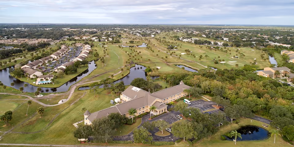 Aerial view of Greenwood Place senior living facility surrounded by green lawns, trees, small ponds, and a golf course. The building is a large, two-story structure with a parking lot and several cars parked. Residential neighborhoods and open land are visible in the background under a cloudy sky.