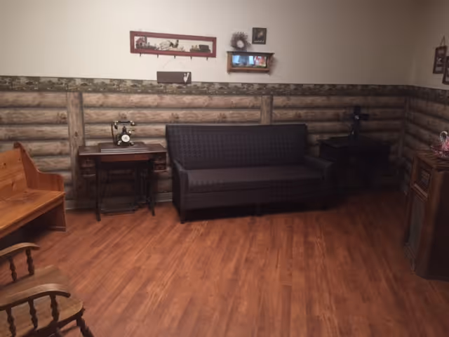 A cozy room with wooden flooring and log cabin style wall paneling. The room contains a dark patterned sofa, a wooden bench, a rocking chair, two small tables with decorative items including an old-fashioned sewing machine, and framed pictures on the wall.