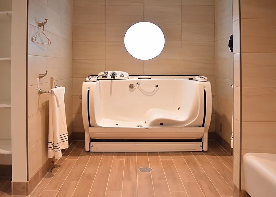 A modern bathroom with a walk-in bathtub featuring safety handles and controls, beige tiled walls, a round window above the tub, a towel hanging on a rack to the left, and wooden-style floor tiles.
