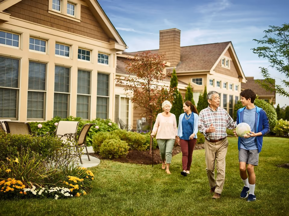 Four people walking outside on a grassy area near a residential building. Two elderly individuals and two younger people are engaged in conversation, with one young person holding a volleyball. The scene is set in a well-maintained garden with shrubs, flowers, and outdoor chairs near the building.