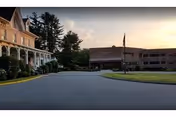 View of the exterior of a healthcare facility at sunset with a paved driveway, a large building on the right, and a historic-style building with a porch on the left, surrounded by trees and greenery.