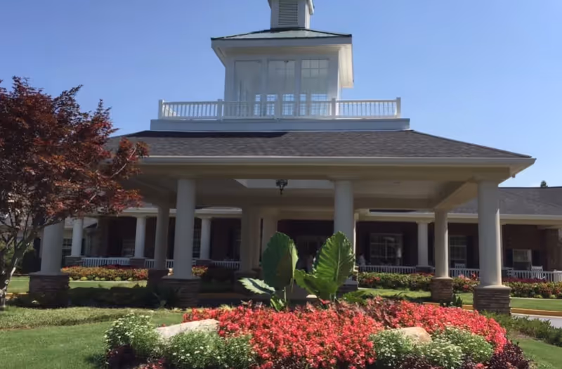Front exterior view of a senior living facility with a covered entrance supported by white columns, a landscaped garden with red and white flowers, green plants, and a tree with red leaves under a clear blue sky.