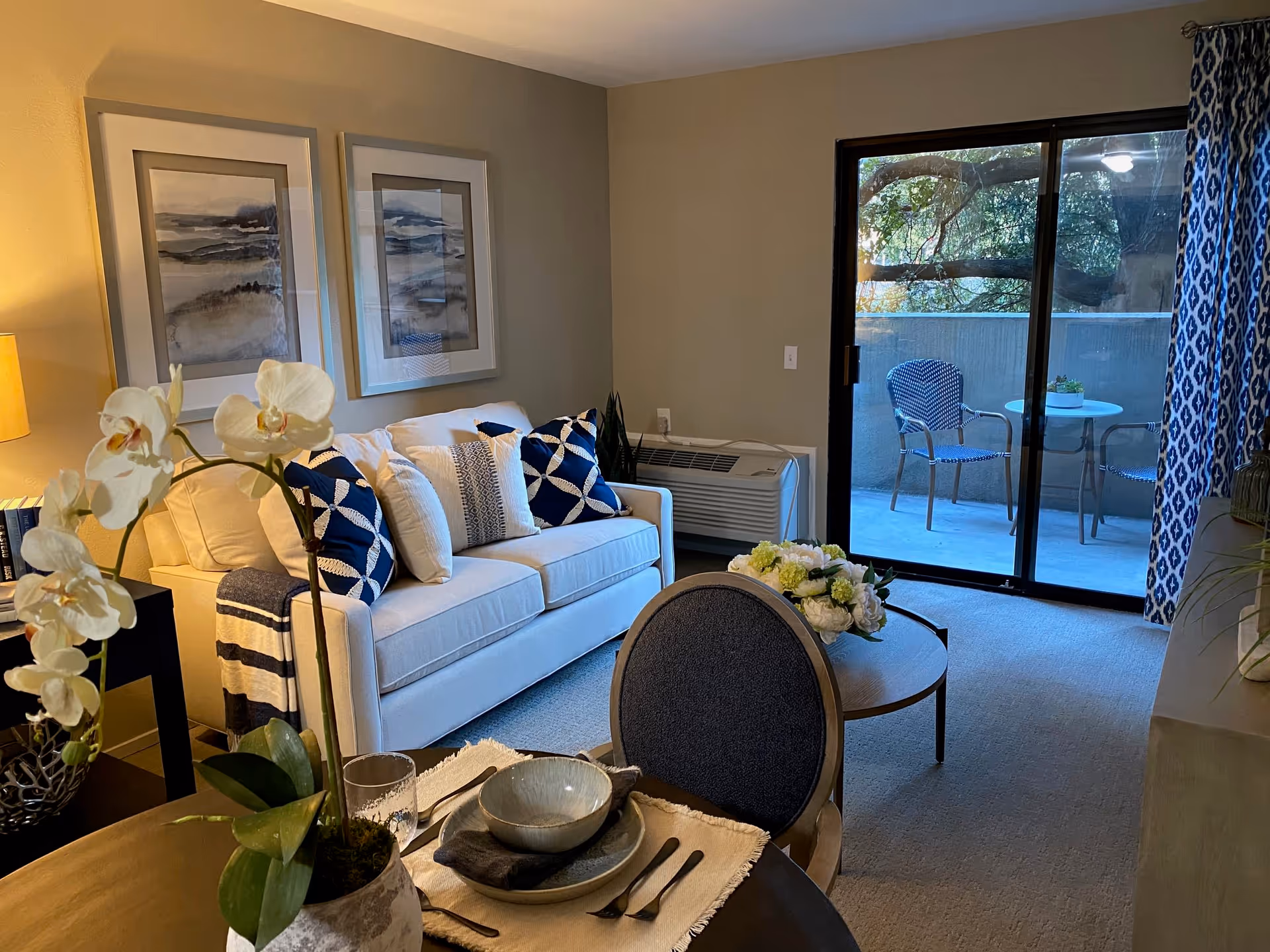 Well-decorated living room with a white sofa, patterned pillows, a coffee table and dining set in the foreground, and a sliding glass door leading to a small patio.