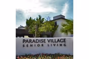 Exterior view of a senior living facility with a sign that reads 'PARADISE VILLAGE SENIOR LIVING' in front of a building surrounded by trees and plants under a partly cloudy sky.