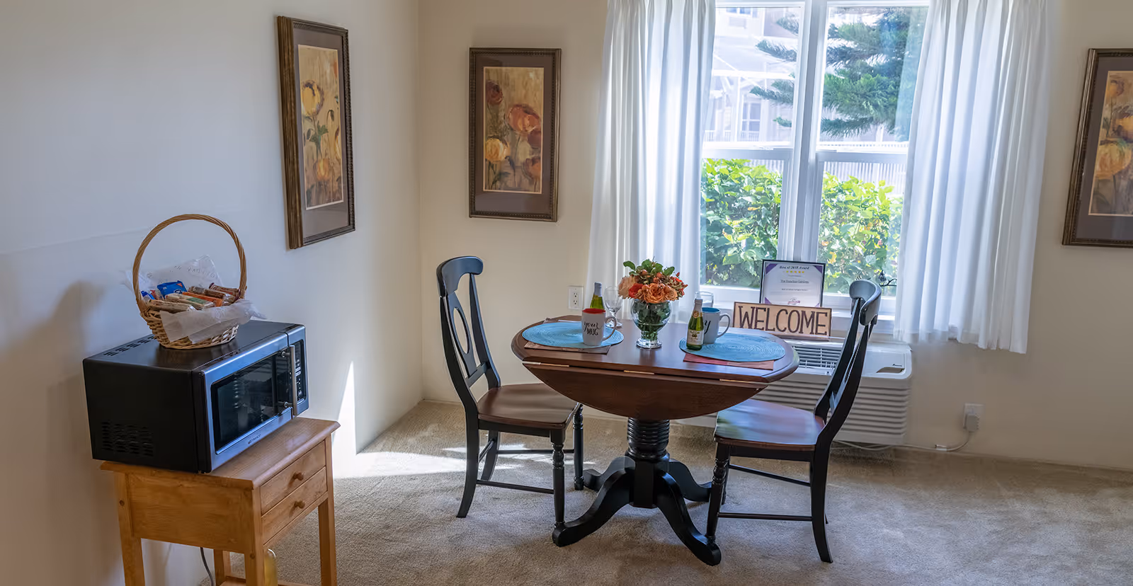 Small dining nook with a round table set for two, two chairs, a microwave on a side table, and a 'WELCOME' sign on the windowsill.