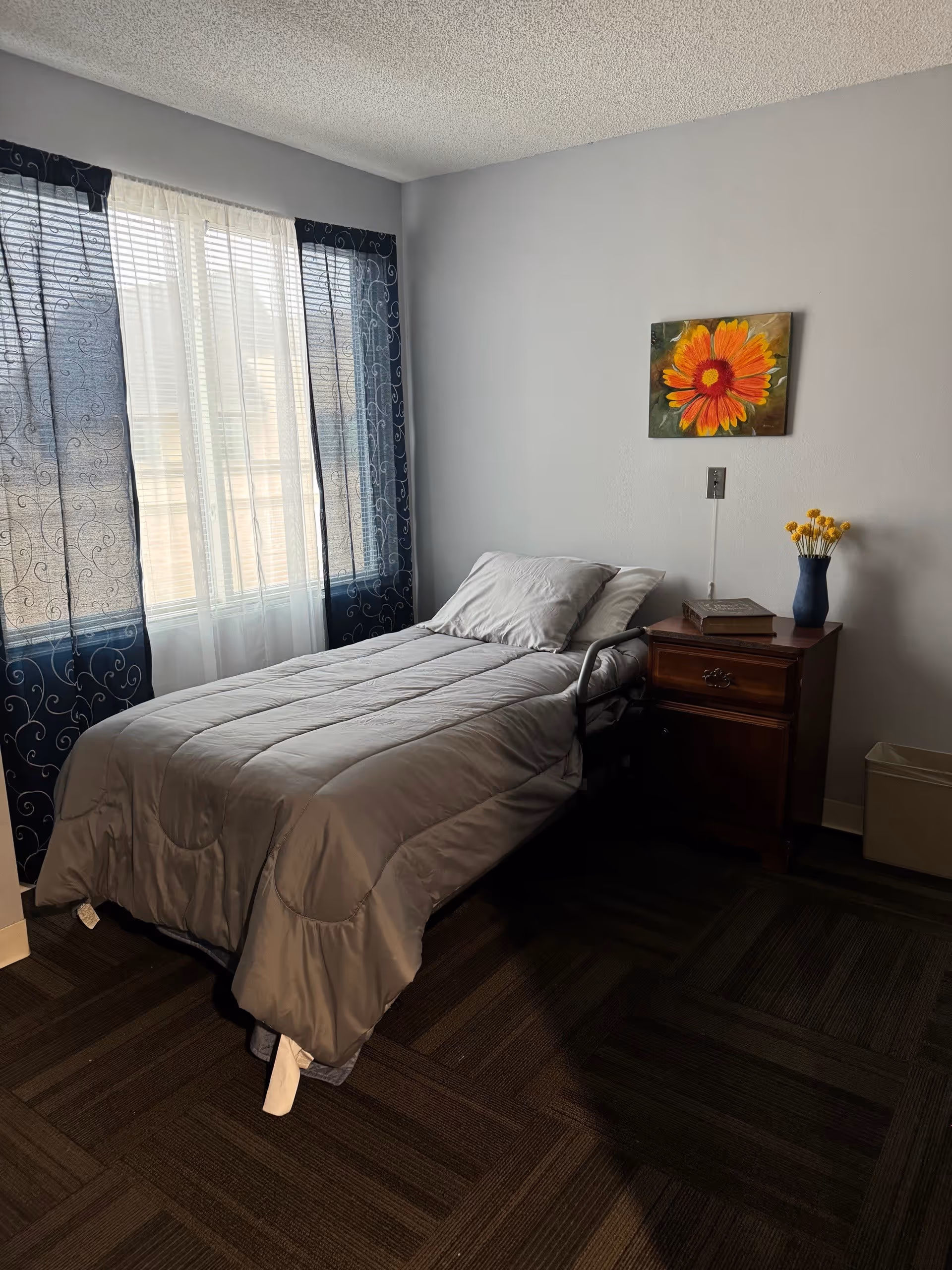 A simple bedroom with a single bed covered in a gray comforter and pillow. Next to the bed is a wooden nightstand with a book and a blue vase holding yellow flowers. A colorful painting of an orange and yellow flower hangs on the light gray wall above the nightstand. The room has a window with sheer white curtains and dark blue patterned drapes, and the floor is covered with dark patterned carpet tiles.