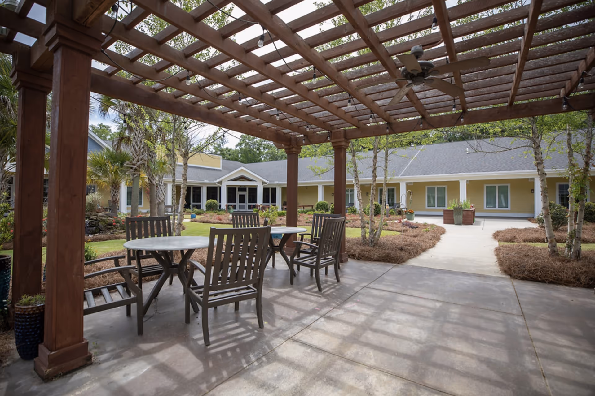 Outdoor patio area with a wooden pergola overhead, string lights, ceiling fan, and several wooden chairs around round tables. The patio overlooks a landscaped garden with trees and shrubs, and a yellow building with white trim in the background.