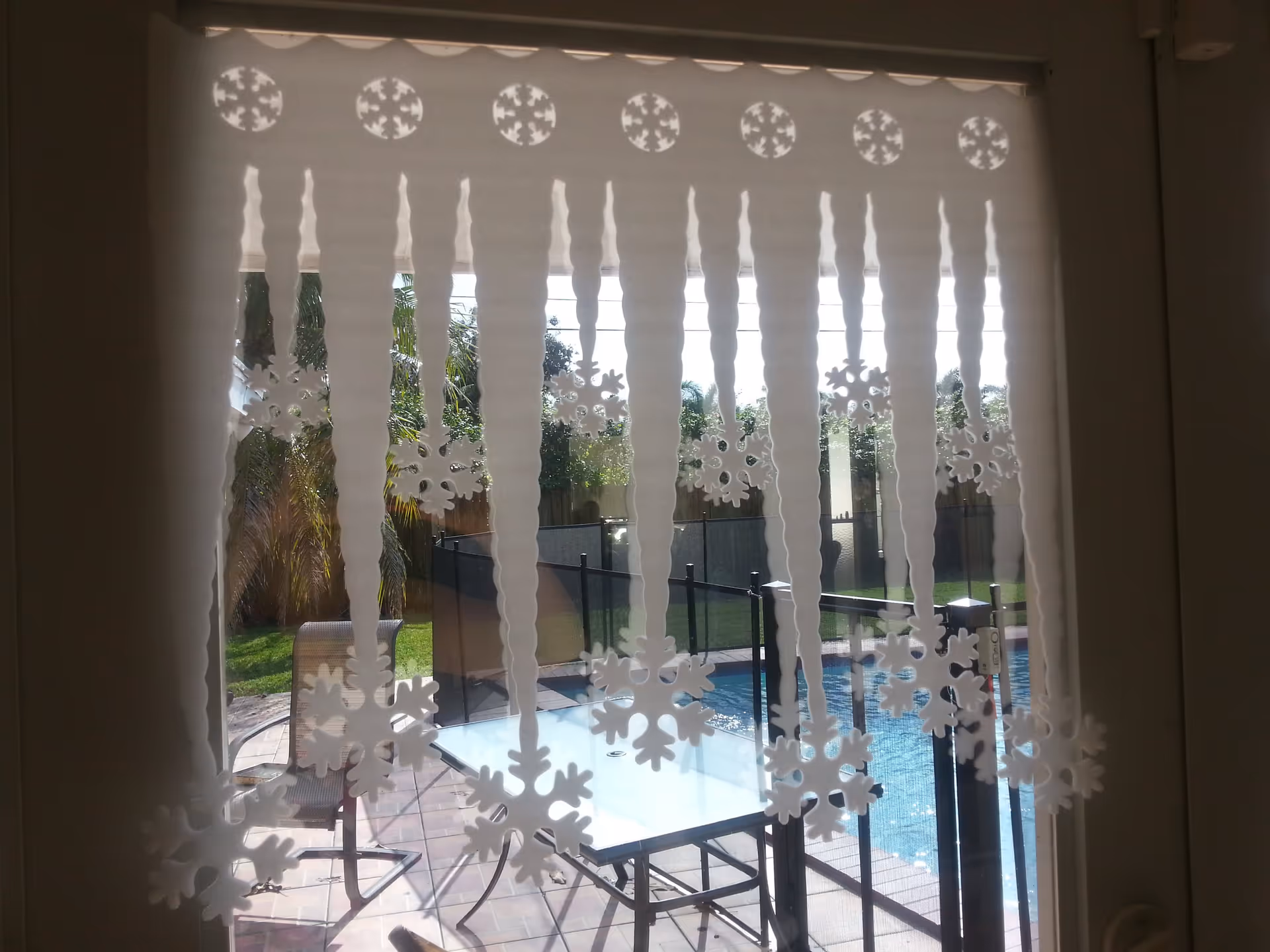 View through a window decorated with hanging snowflake stickers and vertical blinds to a patio table, chairs and a fenced swimming pool.