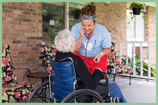 A healthcare worker wearing a light blue shirt and red pants uses a stethoscope to listen to the chest of an elderly woman seated in a wheelchair on a covered porch with floral cushioned chairs and brick walls.