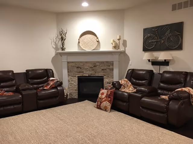 A cozy living room with dark brown leather recliner chairs arranged around a beige woven rug. The room features a white fireplace with a stone surround, decorated with a round woven tray, candles, and a vase with dried branches. A wall-mounted artwork of a bicycle and a double lamp are on the right wall.