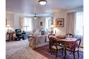 A spacious living and dining area in a retirement center featuring a round dining table with chairs, a sofa, armchairs, lamps, framed artwork on the walls, and large windows with curtains allowing natural light to fill the room.