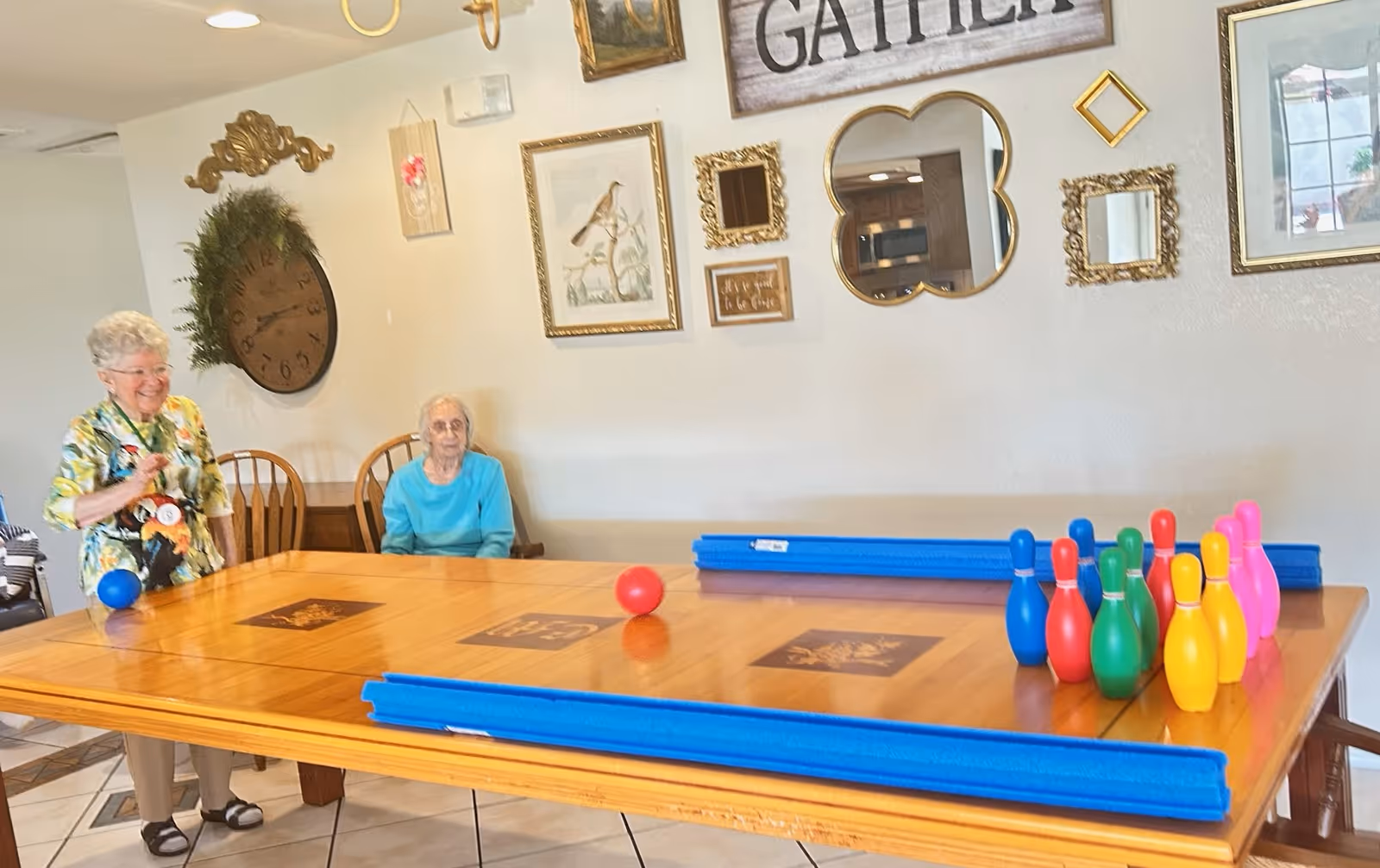 Two elderly women in a room with a wooden table set up for a game of indoor bowling with colorful plastic pins and balls. The wall behind them is decorated with various framed pictures, mirrors, and a large clock. One woman is standing and smiling while holding a blue ball, and the other is seated wearing a turquoise top.