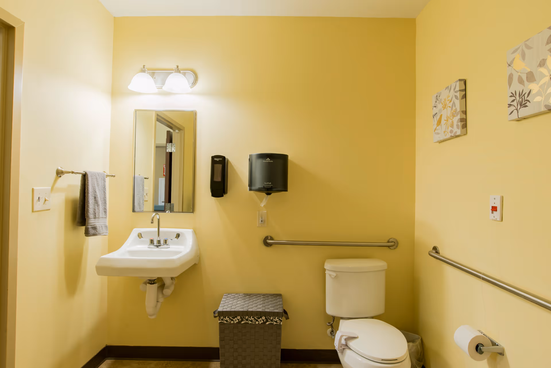 A clean bathroom with yellow walls featuring a white sink with a mirror and light fixture above it, a towel hanging on a rack, a soap dispenser, a paper towel dispenser, a toilet with grab bars on the walls, a wastebasket, and two decorative wall hangings.
