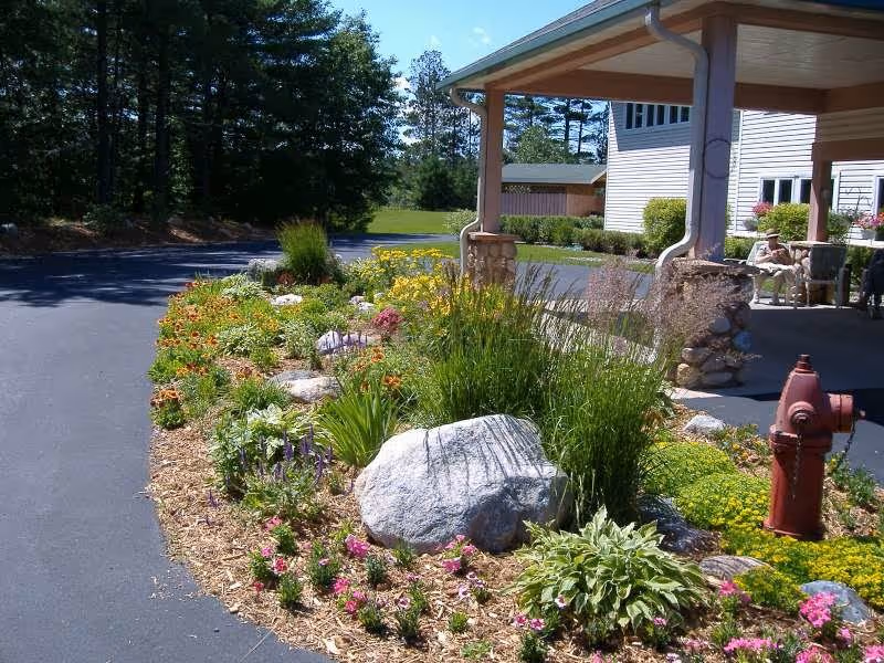 A landscaped garden area with various flowers, plants, and large rocks next to a paved driveway. A red fire hydrant is visible on the right side near the garden. In the background, there is a covered porch attached to a light-colored building with stone pillars and some outdoor seating. Trees and greenery are visible further in the background under a clear blue sky.