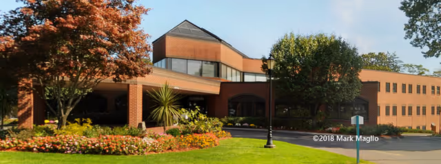 Exterior view of a two-story brick building with large windows, surrounded by well-maintained landscaping including green grass, colorful flower beds, and trees under a clear blue sky.