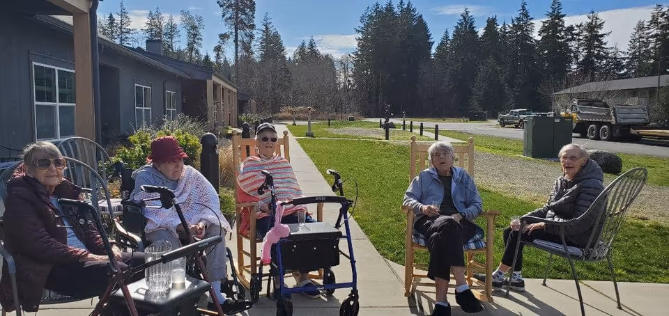 Five elderly women sitting outside on chairs along a sidewalk in front of a building, with walkers nearby. They are enjoying a sunny day with green grass and trees in the background.