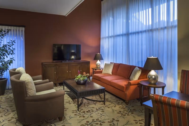 A cozy living room with a red sofa, two brown armchairs, a wooden coffee table with a plant centerpiece, two side tables with lamps, a wooden TV stand with a flat-screen TV, and large windows with sheer white curtains letting in natural light.