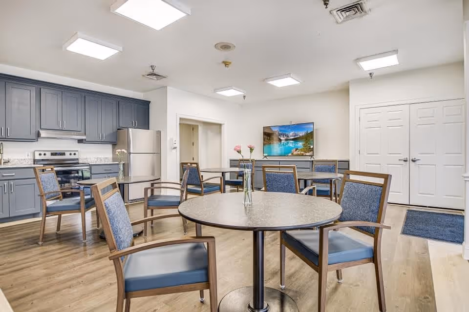 A bright and clean common area in a senior living facility featuring a small kitchen with gray cabinets, stainless steel refrigerator, and stove. Several round tables with blue cushioned chairs are arranged in the room, each table decorated with a vase holding pink flowers. A flat-screen TV is mounted on the wall, and the flooring is light wood. The room has white walls and ceiling with recessed lighting.