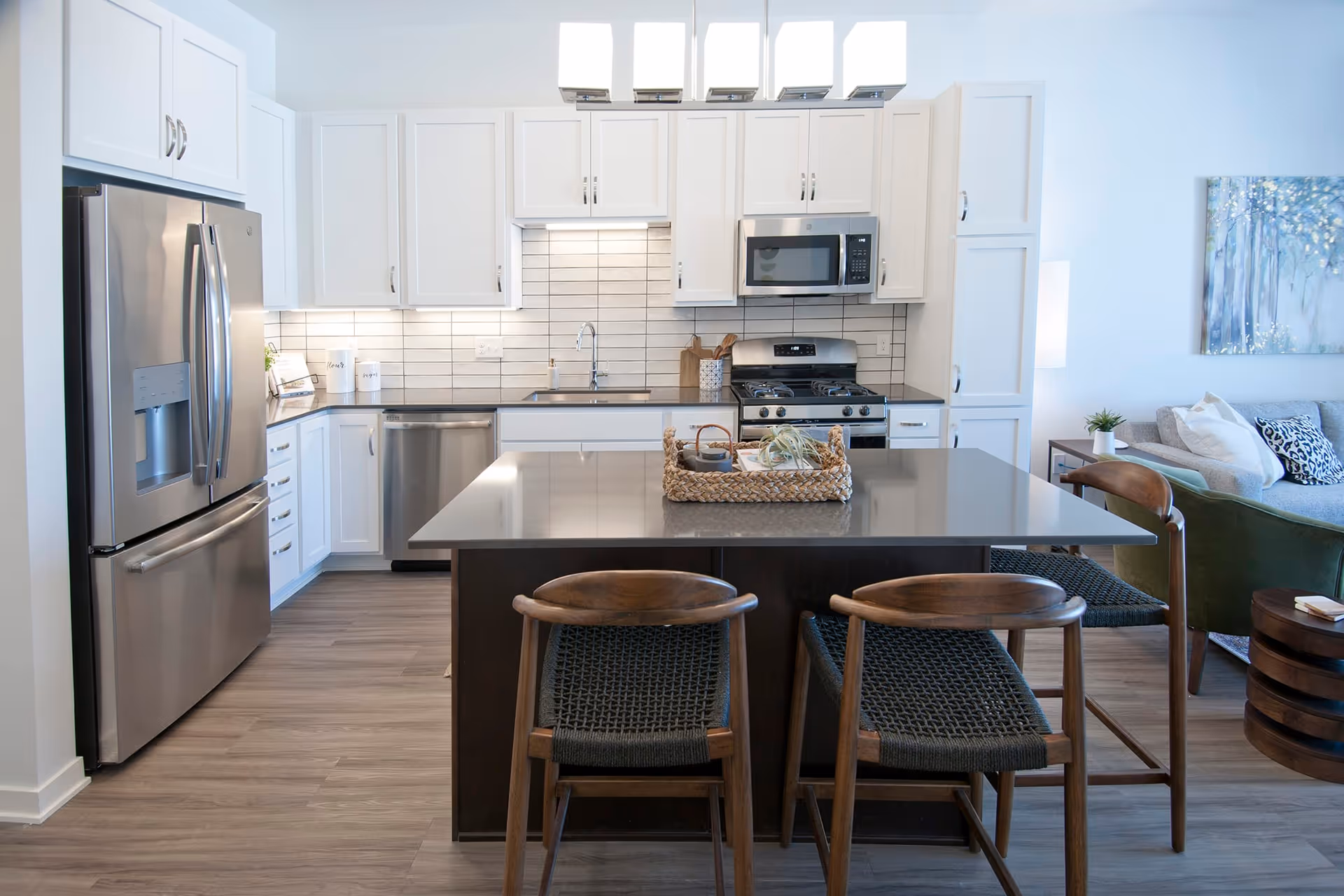 Modern kitchen with white cabinets, stainless steel refrigerator, dishwasher, microwave, and gas stove. A large island with a gray countertop and two wooden chairs with woven seats is in the center. The kitchen opens to a living area with a sofa, armchair, and wall art.