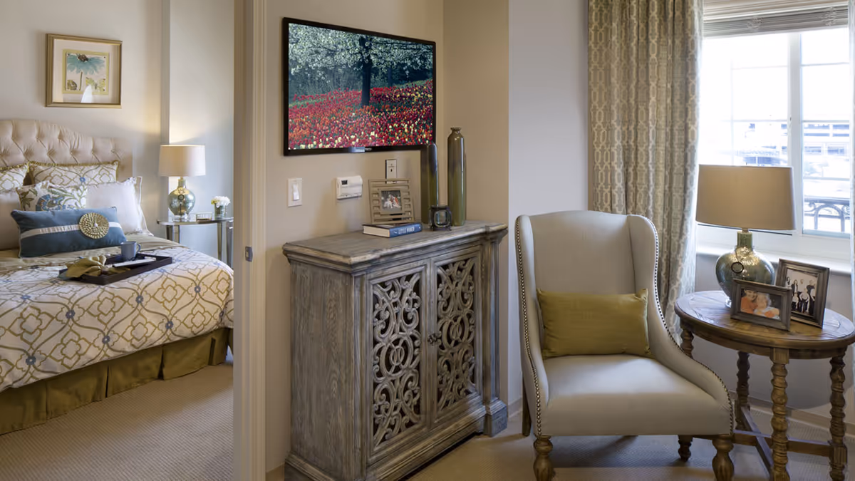 A cozy senior living bedroom with a neatly made bed featuring patterned bedding and decorative pillows. Next to the bed is a nightstand with a lamp and a small flower vase. Adjacent to the bedroom is a sitting area with a cushioned armchair, a wooden side table holding a lamp and framed photos, and a decorative cabinet with books and vases on top. A large window with patterned curtains allows natural light to fill the room.