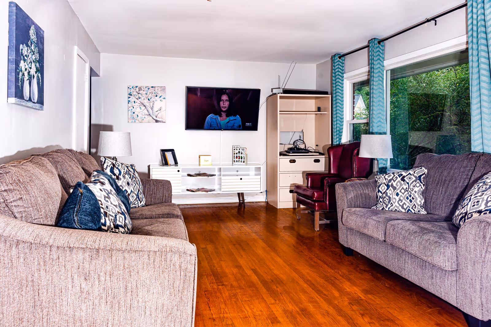 A living room with two sofas facing each other, each with patterned cushions. There is a wooden floor, two table lamps on either side of the sofas, a mounted flat-screen TV on the wall, and a cabinet with electronic devices. Large windows with blue patterned curtains show greenery outside.