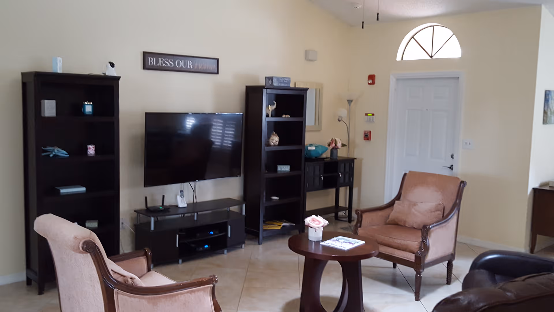 A cozy living room area with two upholstered armchairs and a small round wooden table with a flower vase and a magazine on it. There is a flat-screen TV mounted on the wall between two tall dark wooden bookshelves. A small console table with decorative items and a floor lamp is near a white door with a semi-circular window above it. The walls are light-colored and the floor is tiled.