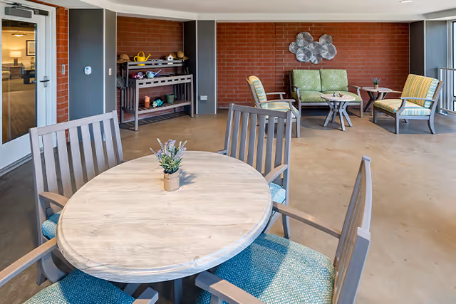 Indoor seating area with a round wooden table and four chairs with blue cushions in the foreground. In the background, there is a brick wall with a green cushioned loveseat, two armchairs with striped cushions, a small round table, and a decorative metal wall art. A shelving unit with gardening tools and pots is against the brick wall. The floor is a smooth concrete surface and there is a glass door leading to another room.