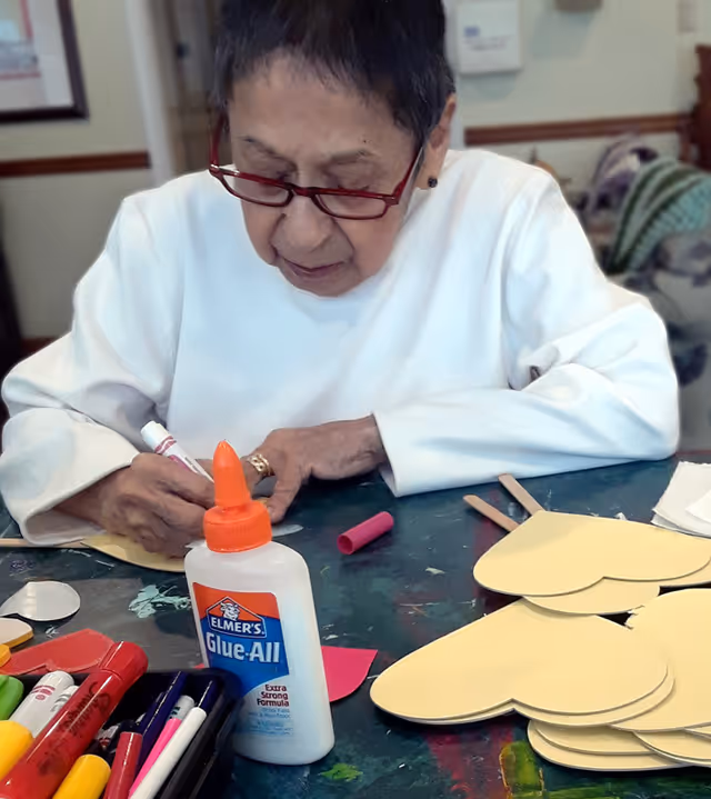 An elderly woman wearing red glasses and a white long-sleeve shirt is sitting at a table engaged in a craft activity. She is writing or drawing on a piece of paper with a marker. On the table in front of her are several yellow heart-shaped cutouts, a bottle of Elmer's Glue-All, various markers, and other craft supplies.