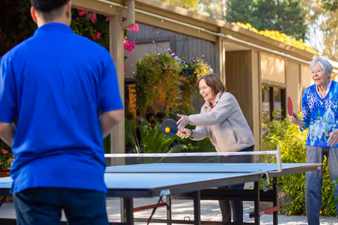 Two elderly women and a man playing table tennis outdoors near a building with plants and flowers around. One woman is actively hitting the ball with a paddle while the other woman watches and smiles. The man is seen from behind wearing a blue shirt.