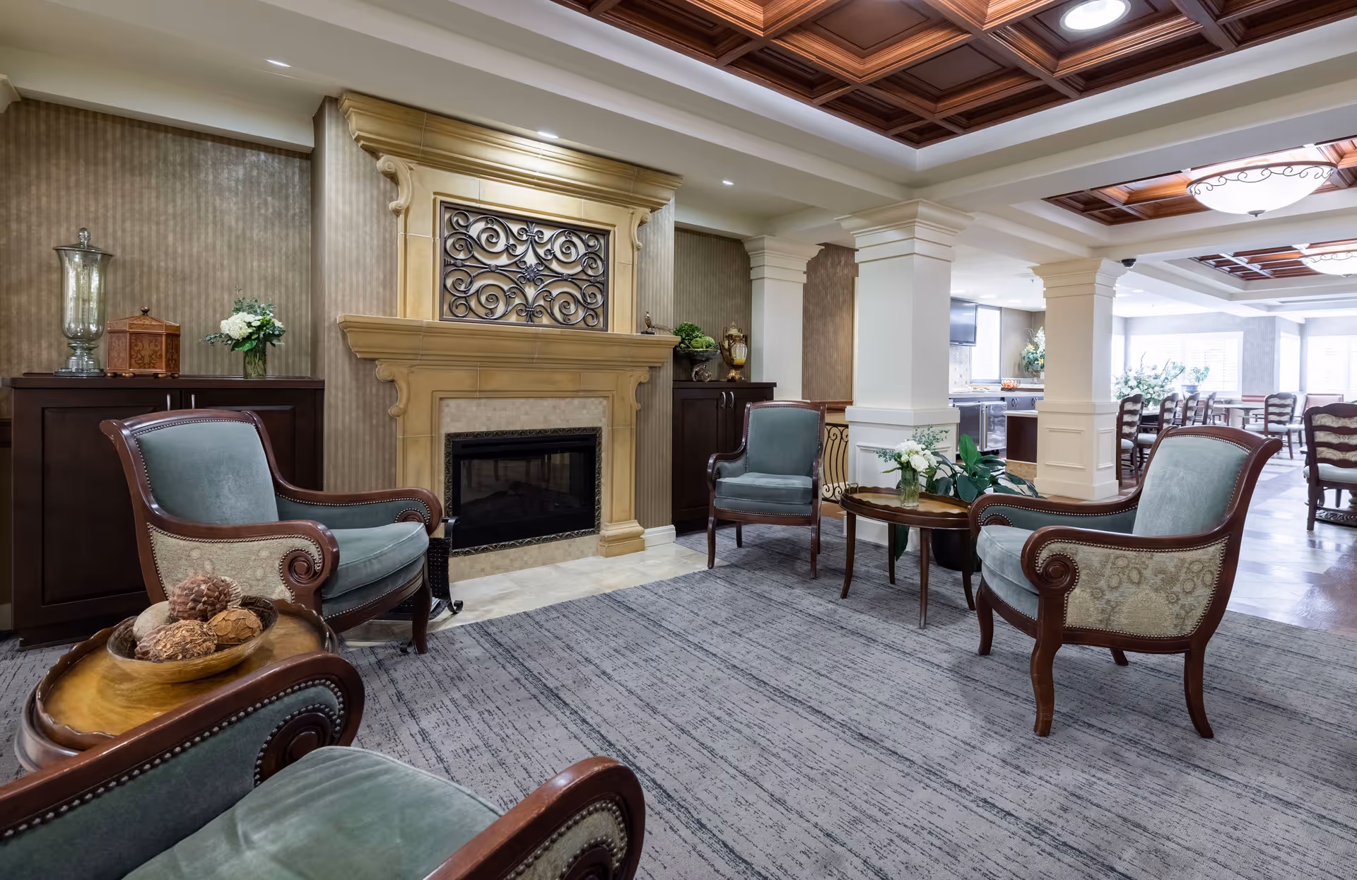 A cozy senior living room area with four upholstered armchairs arranged around a wooden coffee table with a flower arrangement. The room features a decorative fireplace with an ornate metal grate, wooden cabinets on either side, and a patterned area rug. The ceiling has wooden coffered panels with recessed lighting, and in the background, there is a dining area with multiple chairs and tables.