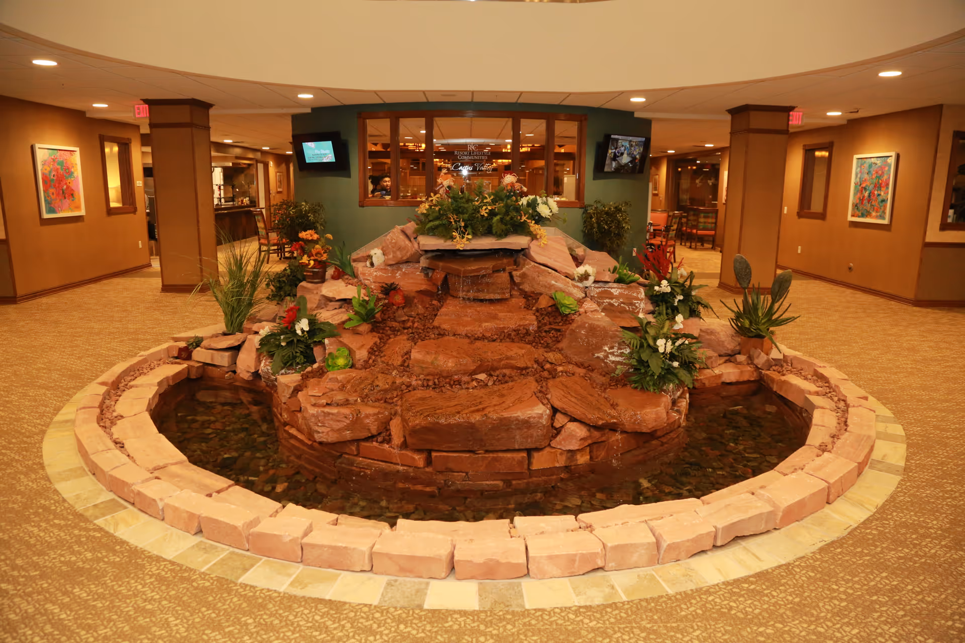 Indoor water feature with red rocks and green plants in the center of a carpeted hallway with brown walls, framed colorful artwork, and two televisions mounted on the walls in a retirement facility.