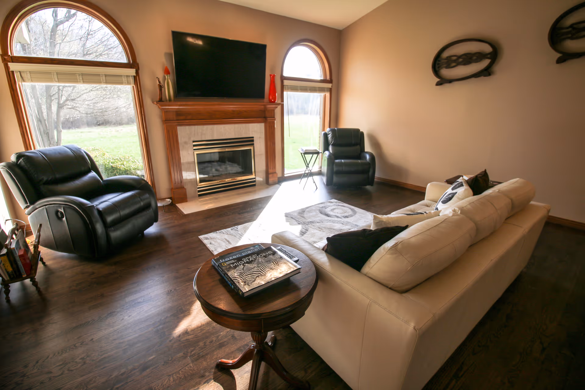 A cozy living room with a beige sofa, two black leather recliners, a wooden round side table with a book on it, a fireplace with a mounted flat-screen TV above it, and two large arched windows letting in natural light. The room has wooden flooring and neutral-colored walls with decorative wall art.