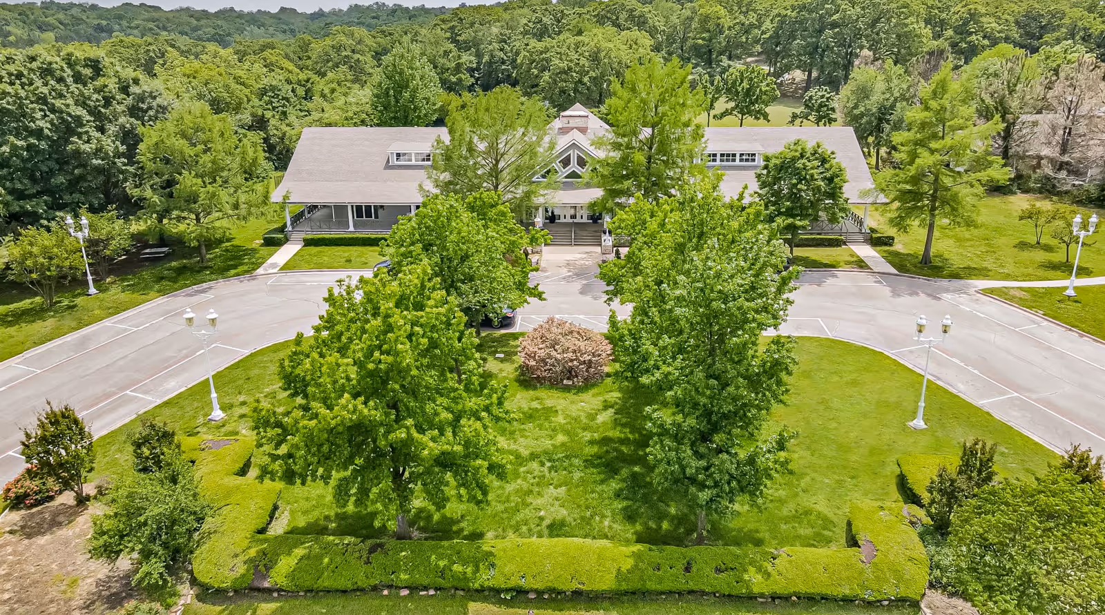 Aerial view of The Homestead of Denison facility showing a large building surrounded by lush green trees and well-maintained lawns. The building has a pitched roof and a central entrance with a driveway and parking area in front.