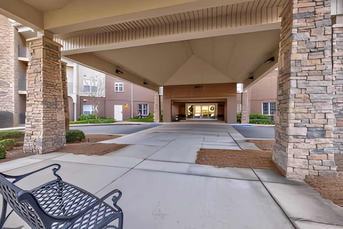 Covered driveway entrance to a building with stone pillars and a metal bench on the left side. The building has a beige exterior with windows and greenery around the entrance area.