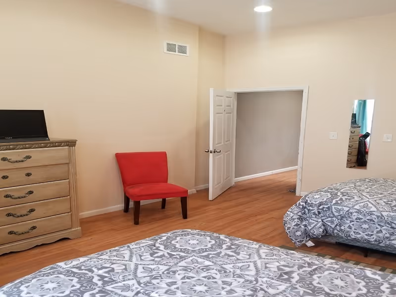 Spacious bedroom with two patterned beds, a red chair, a wooden dresser, and an open door.
