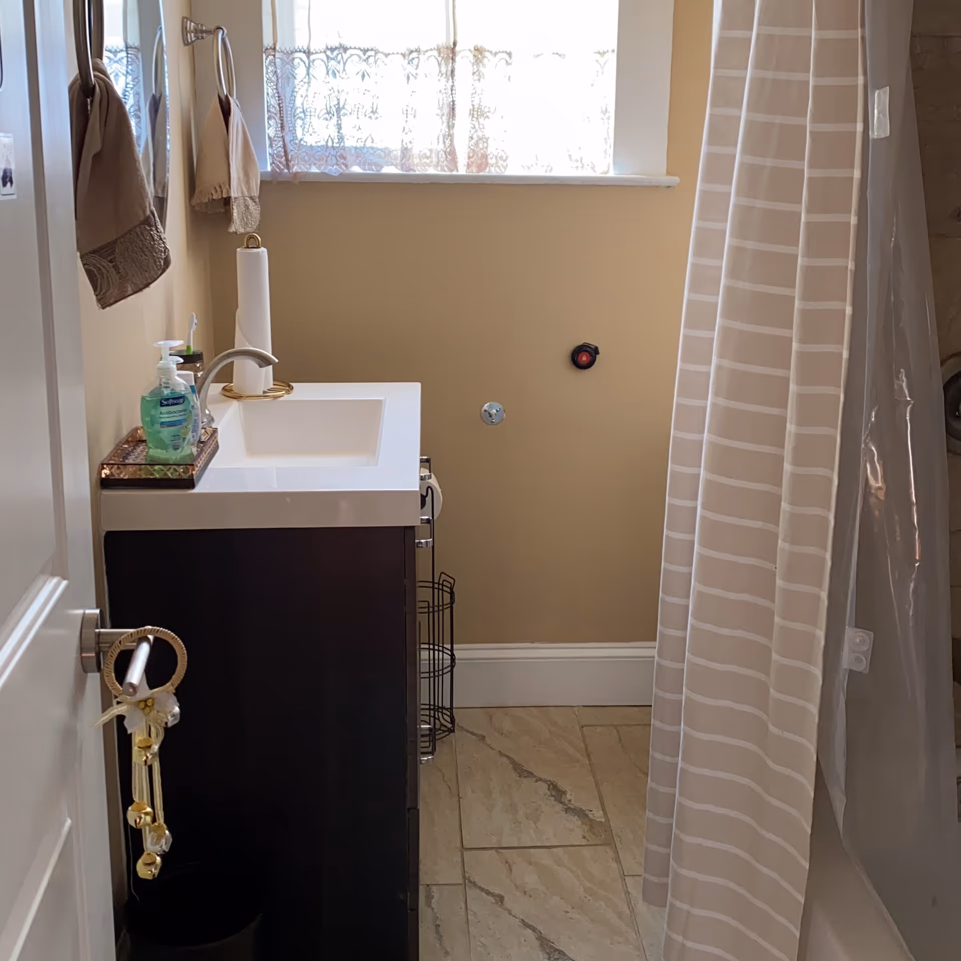 Small bathroom with a sink vanity and soap dispenser, towel hooks, a striped shower curtain, and a window with a lace curtain.