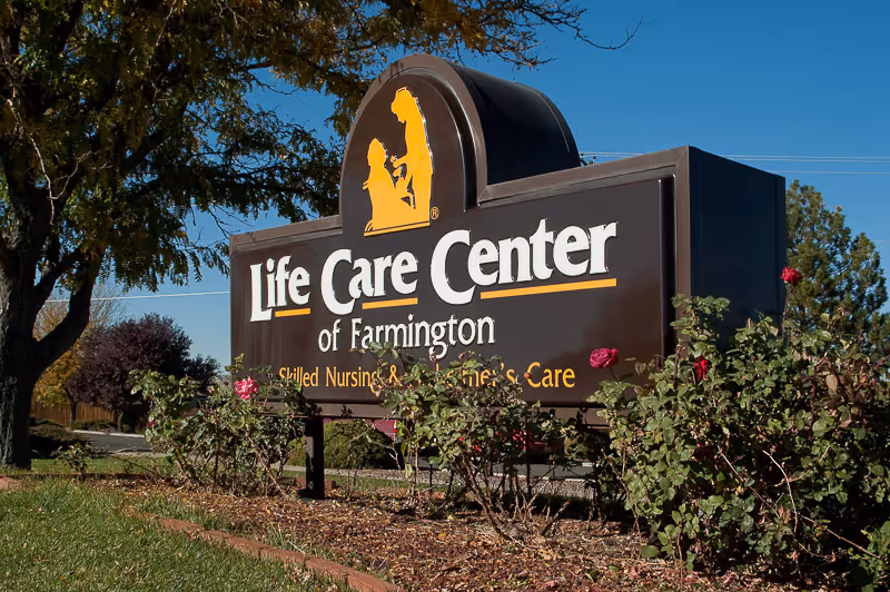 Outdoor sign for Life Care Center of Farmington surrounded by bushes with red flowers, with trees and a clear blue sky in the background.