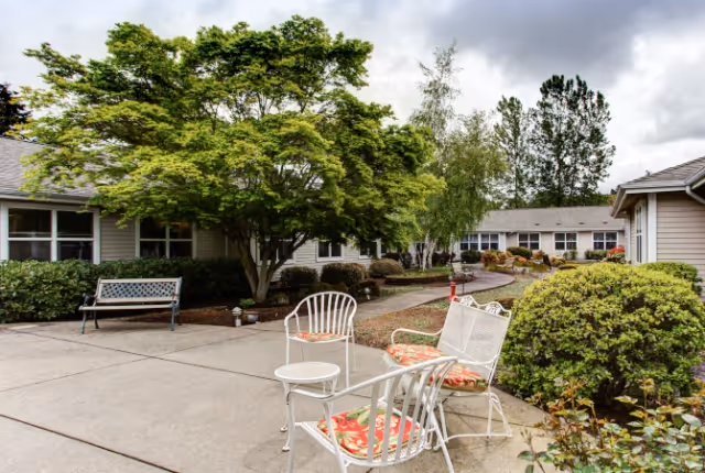 Outdoor patio area at Trustwell Living at Davenport Place featuring white metal chairs with floral cushions, a small round table, a bench, and a large leafy tree surrounded by shrubs and bushes under a cloudy sky.