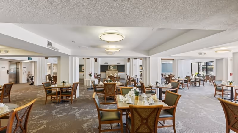 A spacious dining room in a senior living facility with multiple round tables and wooden chairs arranged neatly. Each table is set with white napkins, glassware, and small flower arrangements. The room has large windows allowing natural light to enter, and the walls and ceiling are painted white. There is a carpeted floor with a subtle pattern and ceiling lights providing additional illumination.