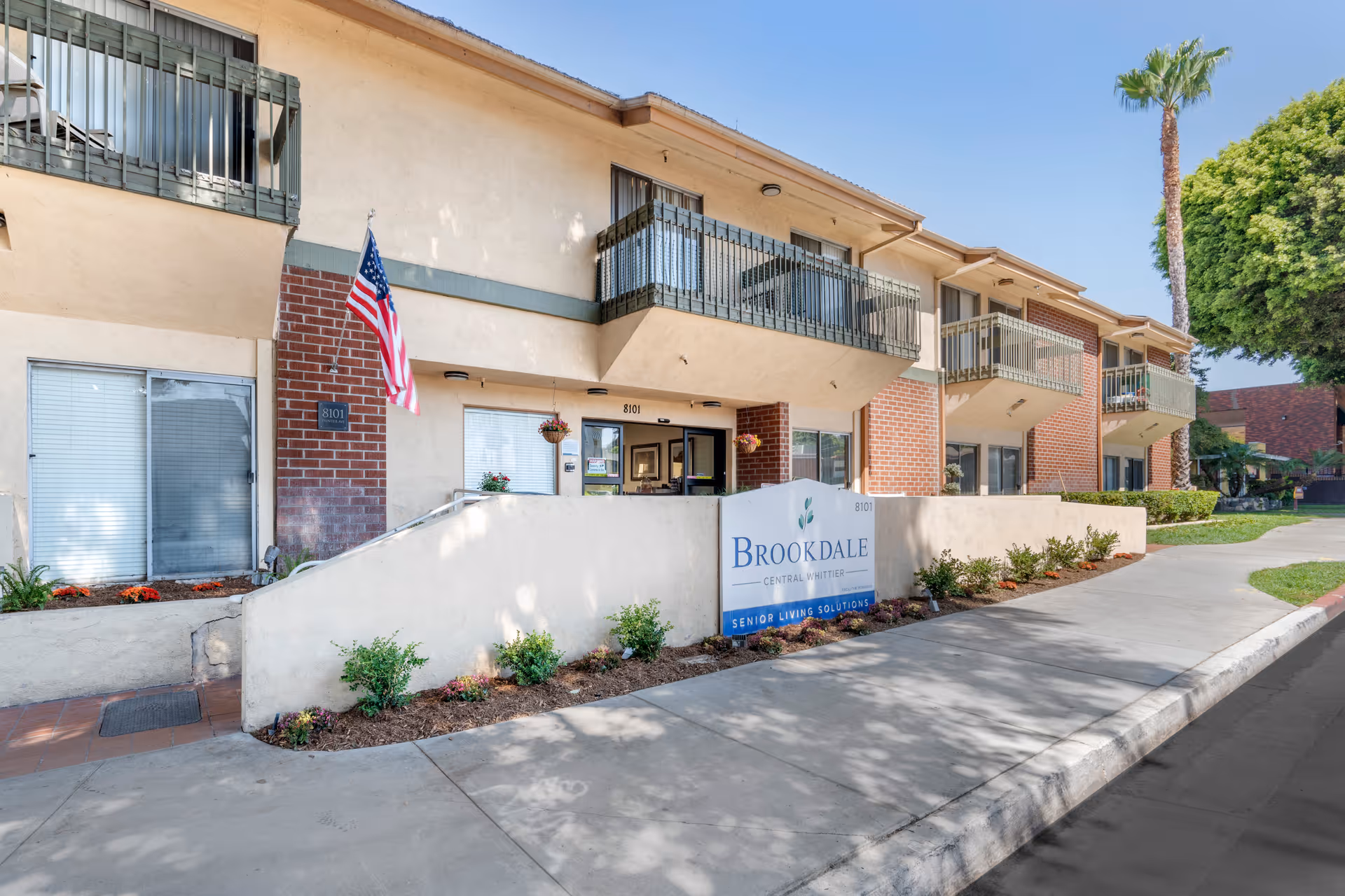 Exterior view of Brookdale Central Whittier senior living facility showing a two-story building with balconies, an American flag, a sign with the facility name, and a sidewalk with landscaping.