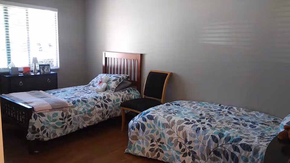 A bedroom with two twin beds covered in matching leaf-patterned bedspreads in shades of blue, gray, and white. There is a wooden nightstand with a lamp, picture frame, and other small items near a window with blinds. A black chair with wooden legs is placed between the beds. The room has light-colored walls and wooden flooring.