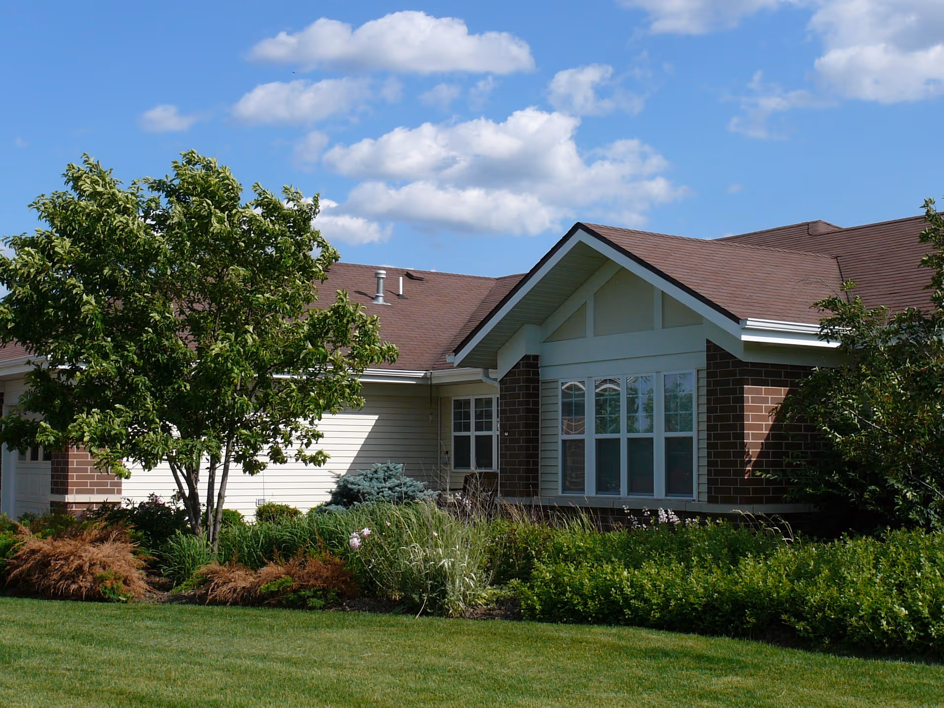 Exterior view of a single-story residential building with a brown roof, brick and light-colored siding, surrounded by green shrubs, trees, and a well-maintained lawn under a blue sky with scattered clouds.