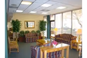 A cozy living room area with striped upholstered sofas and wooden tables. The room has large windows letting in natural light, potted plants, a mirror on the wall, and a ceiling with fluorescent lights.