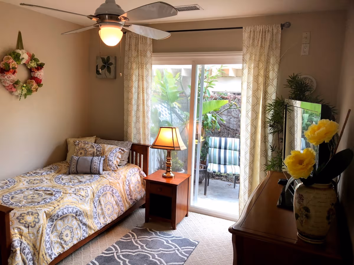 A cozy bedroom with a single bed covered in a yellow and gray patterned comforter and pillows. A wooden nightstand with a lit lamp is beside the bed. There is a ceiling fan with a light above. Sliding glass doors with cream-colored curtains lead to a small outdoor patio with two striped chairs and green plants. A wooden dresser with a vase of yellow flowers and a TV is on the right side of the room. A floral wreath hangs on the wall above the bed.