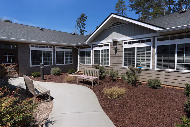 Courtyard with a curved concrete walkway, benches, and landscaped mulch beds in front of a single-story building.