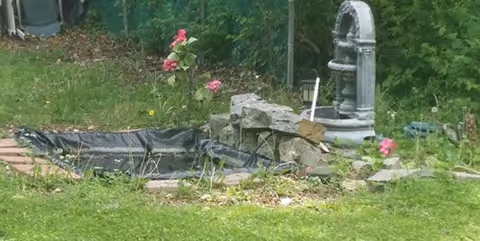 A small outdoor garden area with a partially covered pond, surrounded by grass and some pink flowers. There is a decorative stone water pump feature next to the pond and some greenery in the background.