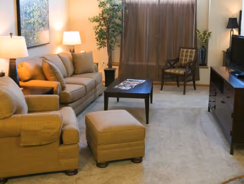 A cozy living room with beige upholstered furniture including a sofa, armchair, and ottoman. There is a dark wooden coffee table with magazines on it, a wooden TV stand with a flat-screen TV, a wooden chair with a cushion, two table lamps, a tall potted plant, and a large window covered with brown curtains.