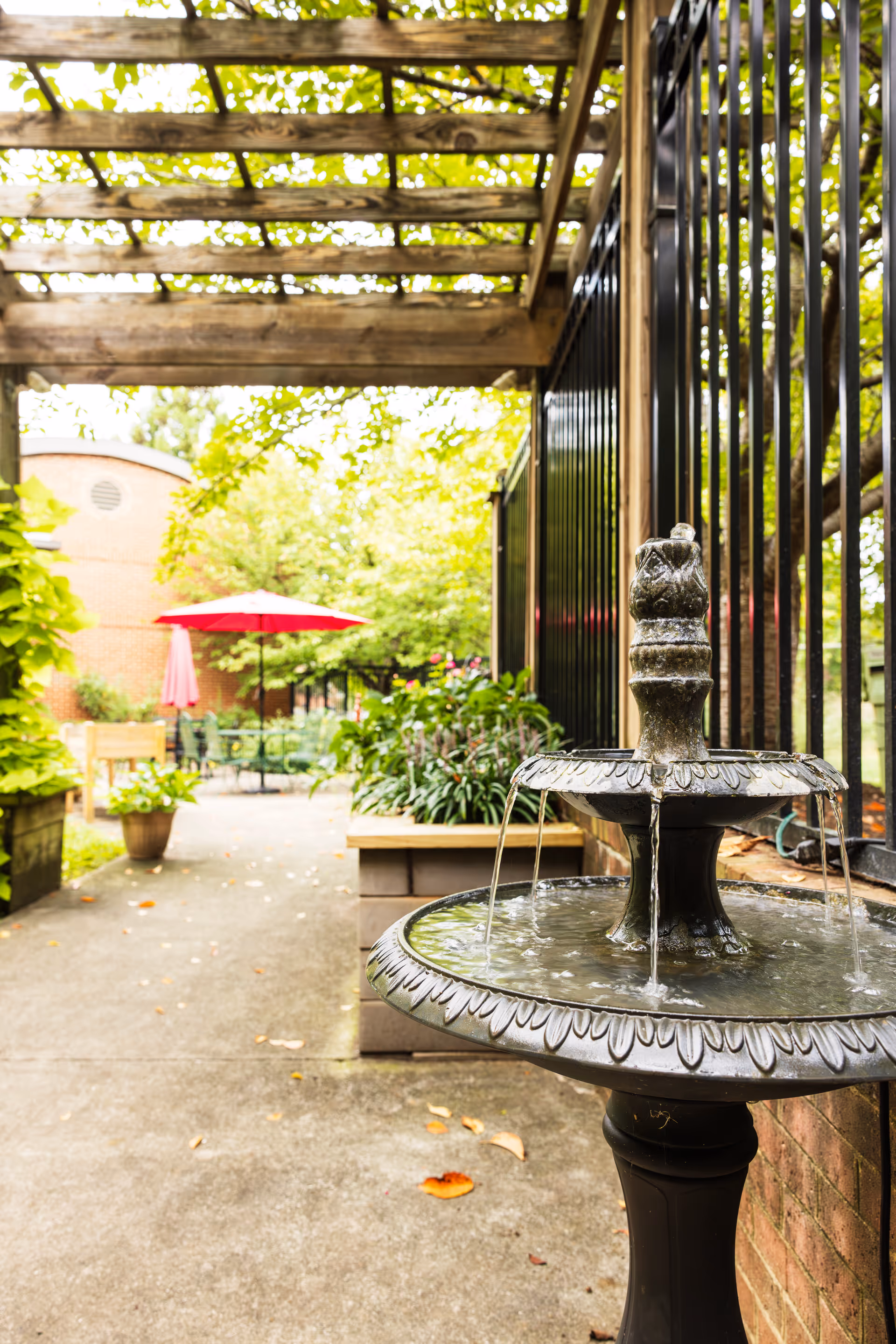 Outdoor garden area with a decorative two-tier water fountain in the foreground, a wooden pergola overhead, potted plants, and a seating area with tables and chairs under red umbrellas in the background.