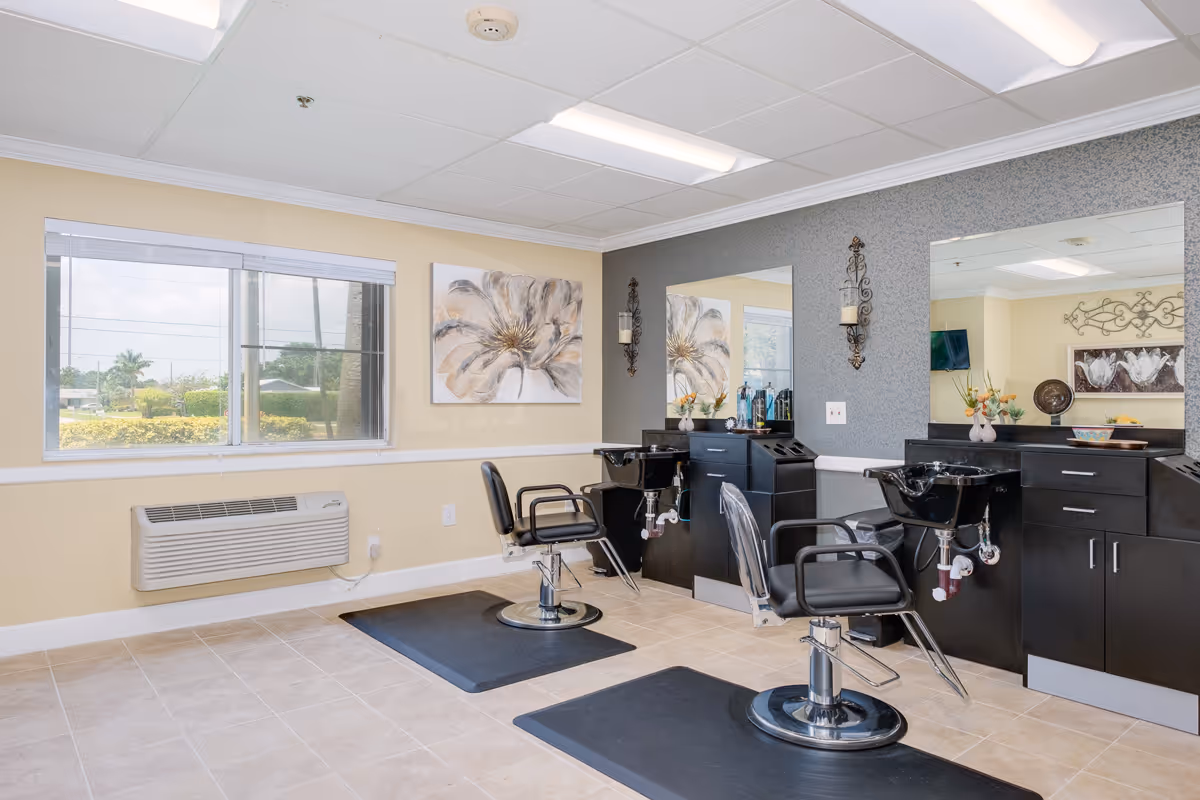 Interior view of a hair salon area in a senior living facility with two black salon chairs in front of large mirrors, black cabinetry with sinks, and decorative wall art. A window shows greenery outside, and the room has tiled floors and fluorescent ceiling lights.