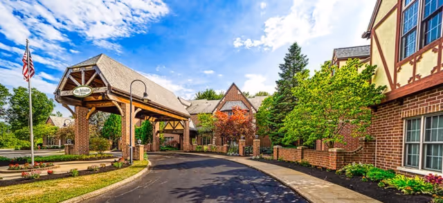 Exterior view of Saint Therese of Westlake facility showing a brick building with Tudor-style architectural elements, a covered entrance with a sign that reads 'The Residence,' landscaped greenery, trees, and a clear blue sky with some clouds.